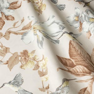 May include: Cream-colored fabric with a floral and hummingbird pattern. The design features large white and gray flowers, brown leaves, and small yellow flowers. Hummingbirds are depicted in flight, adding a touch of nature to the fabric.