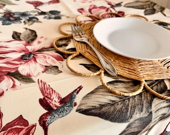May include: A white ceramic plate with a woven placemat and a silver fork on a cream tablecloth with a floral pattern featuring pink flowers and hummingbirds.