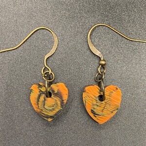May include: A pair of earrings with a heart-shaped design. The hearts are made of orange and black patterned clay and have a gold-toned hook.