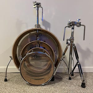 May include: A three-piece drum set with a bass drum, a snare drum, and a tom-tom. The drums are all made of wood and have a natural finish. The bass drum has a chrome hoop and a chrome lug. The snare drum has a chrome hoop and a chrome lug. The tom-tom has a chrome hoop and a chrome lug. The drums are all set up on a stand with a chrome finish.