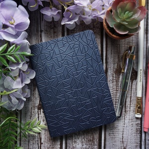 May include: A dark blue notebook with a geometric pattern embossed on the cover. The notebook is lying on a wooden surface with a succulent plant, pens, and flowers.