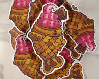 May include: A stack of colorful stickers featuring a cartoon illustration of a taiyaki, a Japanese fish-shaped pastry, filled with pink ice cream and topped with chocolate sprinkles and a chocolate bar.