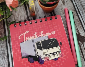 May include: A square spiral-bound notebook with a red cover featuring a white grid pattern, a graphic of a white box truck, and the text Truck-kun with a small heart. The notebook is accompanied by a pink pen and a light green pen.