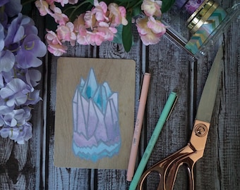 May include: A wooden card with a hand-painted watercolor illustration of a crystal in shades of pink, blue, and purple. The card is surrounded by pink flowers, green leaves, and two pens.