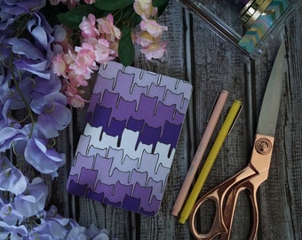 May include: A purple and white notebook with a repeating pattern of cats. The notebook is on a wooden surface with pink flowers and a pair of gold scissors.