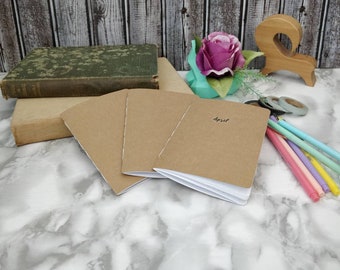May include: Three brown notebooks with white pages, the top notebook has the word "April" written on the front cover. The notebooks are on a white marble surface with a pink flower, a wooden figure eight, and colorful pens.