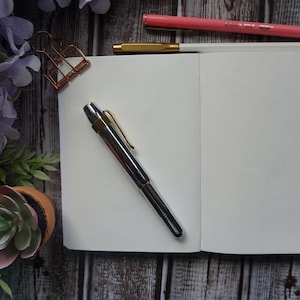 May include: A white notebook with a silver pen on a wooden surface. The notebook is open to a blank page. There are flowers and a potted succulent in the background.