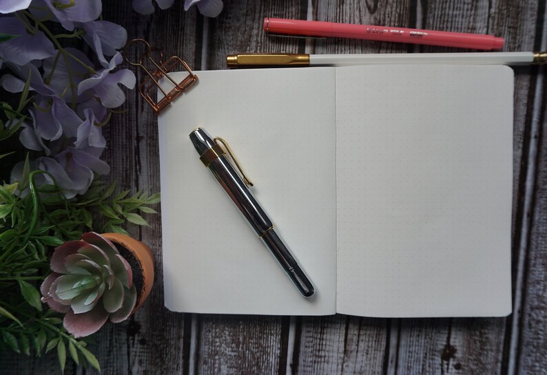 May include: A white notebook with a silver pen on a wooden surface. The notebook is open to a blank page. There are flowers and a potted succulent in the background.