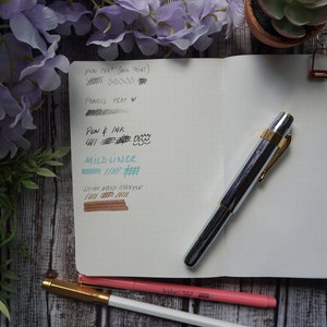 May include: A silver and gold fountain pen with a black ink sample on a white notebook page. The notebook page also has a gray pencil test, a blue ink sample, and a brown ink sample. The notebook is open and has a gold binder clip.