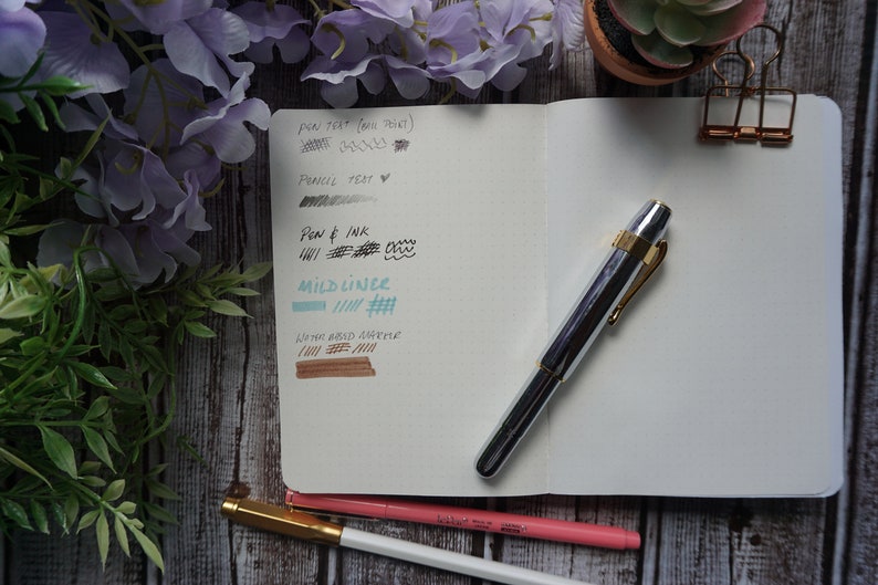 May include: A silver and gold fountain pen with a black ink sample on a white notebook page. The notebook page also has a gray pencil test, a blue ink sample, and a brown ink sample. The notebook is open and has a gold binder clip.