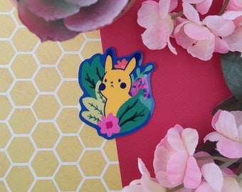 May include: A yellow cartoon character with a blue background and green leaves. The character has a pink flower behind it. The sticker is on a yellow and white honeycomb background.
