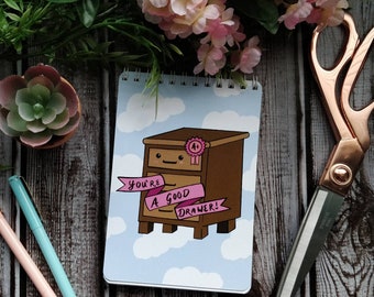 May include: A blue and white notepad with a cartoon illustration of a brown dresser with a pink ribbon and the text "You're a good drawer!"