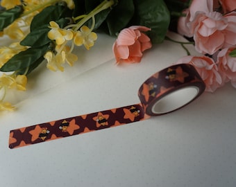 May include: A roll of washi tape with a repeating pattern of orange stars and brown bees. The tape is on a white surface with small brown dots.