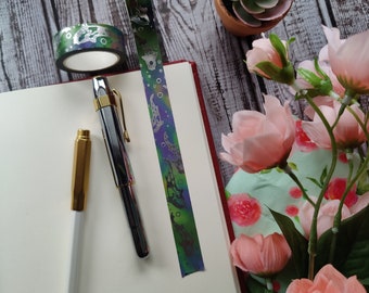 May include: A white notebook with a silver and green washi tape with a pattern of fish and bubbles. Two pens, one gold and one silver, are on the notebook. A small potted succulent and pink flowers are in the background.