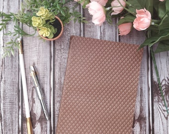 May include: A brown leather notebook with a quilted texture sits on a wooden surface. A white pencil and a silver pen are next to the notebook. A small potted succulent plant and pink flowers are in the background.