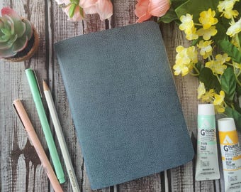 May include: A blue leather-bound journal with a textured cover, surrounded by pens, pencils, and paint tubes. The paint tubes are labeled "Gouache" and are in shades of green and yellow.