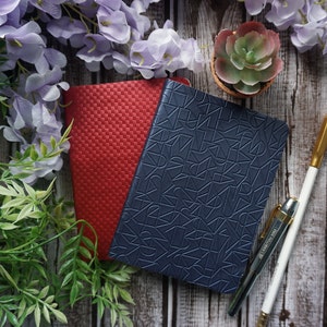 Pu&ograve; includere: Due quaderni, uno rosso e uno blu, con un motivo geometrico sul quaderno blu. Una penna dorata e una matita bianca sono su una superficie di legno con una piccola pianta grassa in vaso e fiori viola.