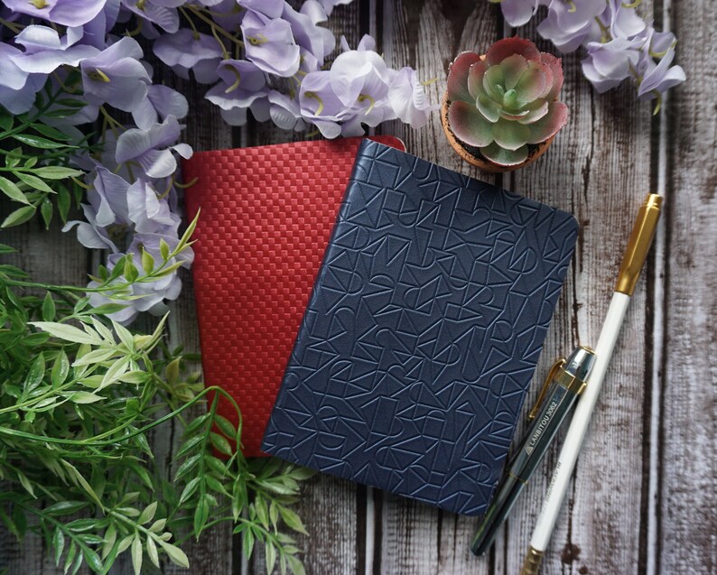 Pu&ograve; includere: Due quaderni, uno rosso e uno blu, con un motivo geometrico sul quaderno blu. Una penna dorata e una matita bianca sono su una superficie di legno con una piccola pianta grassa in vaso e fiori viola.