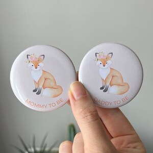 May include: Two round buttons with a pink background and a cartoon fox wearing a flower crown. The text "Mommy to be" is on one button and "Daddy to be" is on the other.