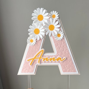 May include: Pink letter A cake topper with a white border and a floral pattern. The letter is decorated with white paper daisies with yellow centers. The name "Anna" is written in orange script on the letter.