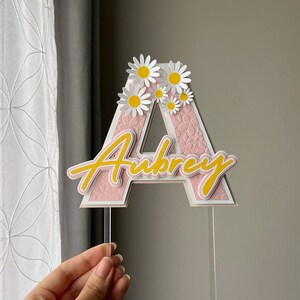 May include: Pink and white letter A cake topper with the name Aubrey written in yellow script. The letter A is decorated with white daisies.