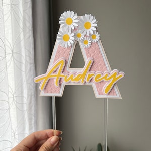 May include: Pink letter A cake topper with the name "Audrey" written in yellow script and white daisy flowers on top. The topper is on two clear plastic sticks.