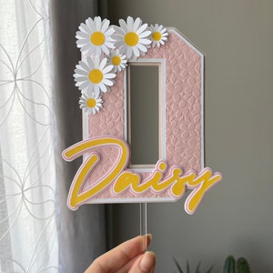 May include: Pink and white cake topper with the letter "D" decorated with white daisies and the word "Daisy" in yellow and pink lettering.