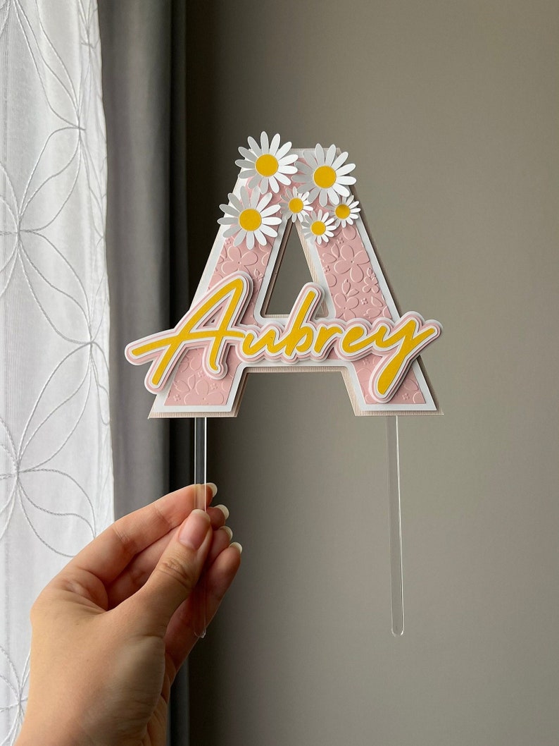 May include: Pink and white letter A cake topper with the name Aubrey written in yellow script. The letter A is decorated with white daisies.