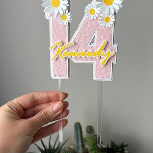 May include: Pink and white cake topper with the number 14 and the name "Kennedy" in yellow script. The number is decorated with white daisies.