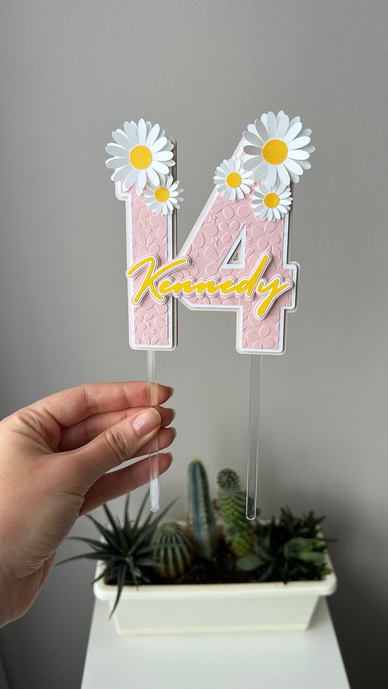 May include: Pink and white cake topper with the number 14 and the name "Kennedy" in yellow script. The number is decorated with white daisies.