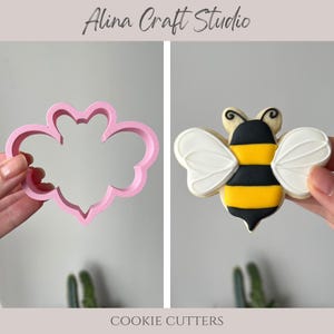 May include: A pink plastic bee-shaped cookie cutter next to a decorated bee-shaped cookie. The cookie has yellow, black, and white icing. The text "Alina Craft Studio" and "COOKIE CUTTERS" are visible.