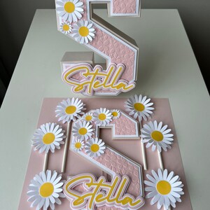 May include: A pink and white 3D number two cake topper with a daisy design and the name "Stella" written in yellow. The topper is surrounded by smaller daisy cake toppers on sticks.