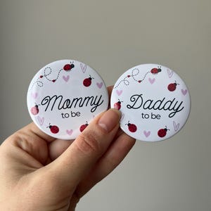 May include: Two white round buttons with the text "Mommy to be" and "Daddy to be" in elegant script. Each button features ladybug and heart illustrations. The buttons are held in a hand.