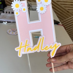 May include: Pink letter H with a white outline decorated with white paper daisies. The letter is attached to a clear plastic stick and has the name "Hadley" written in yellow below it.
