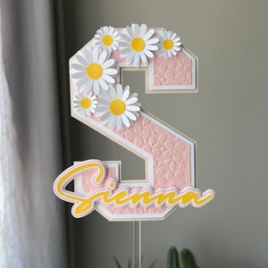 May include: A pink and white cake topper with the letter "S" decorated with white daisies and the name "Sienna" in yellow. The topper is on a clear plastic stick.