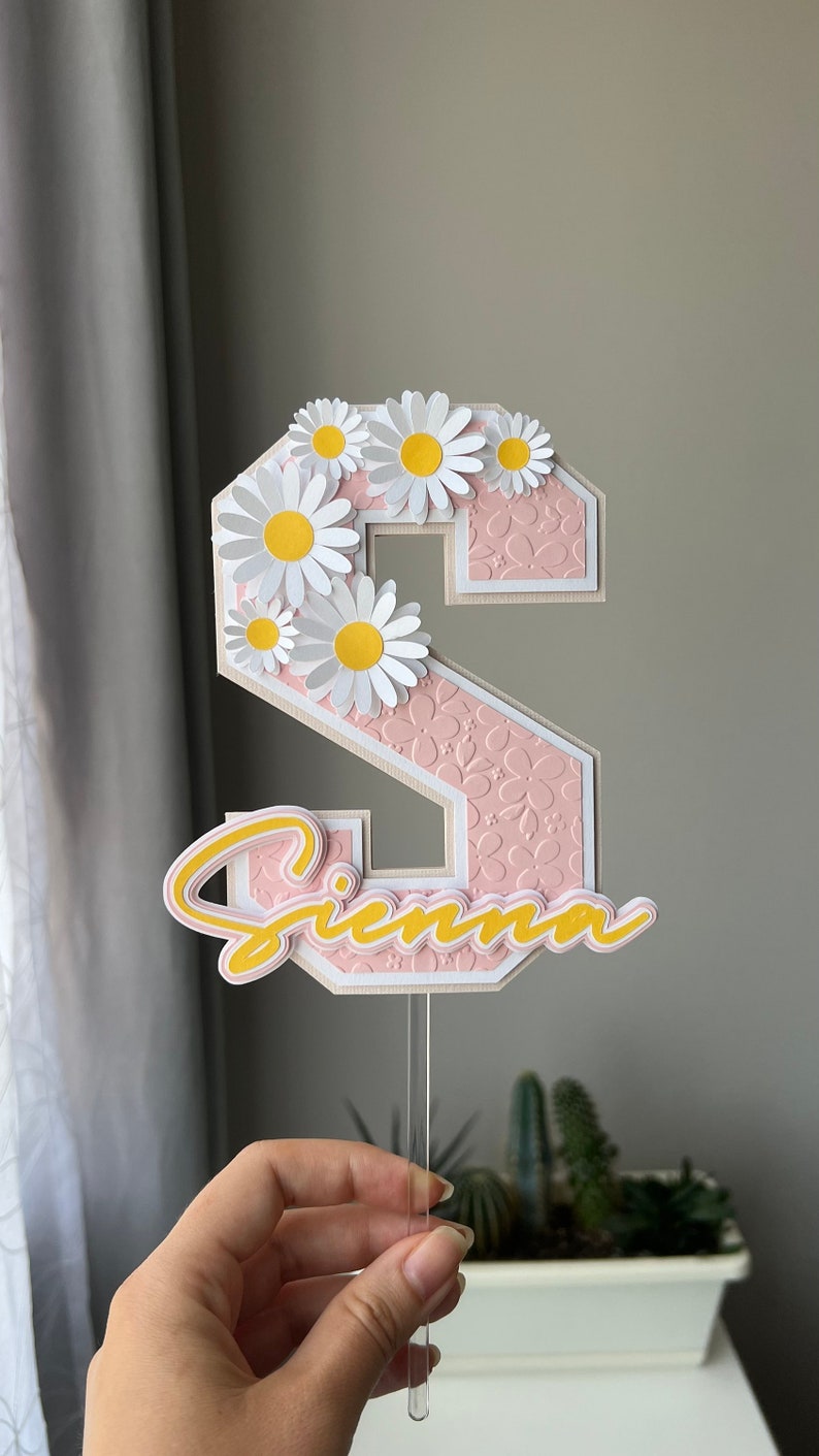 May include: A pink and white cake topper with the letter "S" decorated with white daisies and the name "Sienna" in yellow. The topper is on a clear plastic stick.