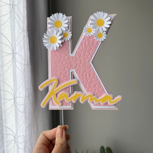 May include: Pink letter K with white outline and a floral pattern. The letter is topped with white paper daisies with yellow centers. The word "Karma" is written in yellow script below the letter.