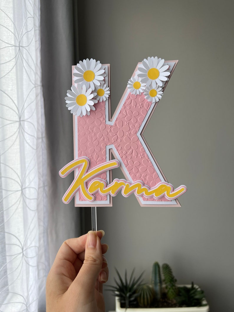 May include: Pink letter K with white outline and a floral pattern. The letter is topped with white paper daisies with yellow centers. The word "Karma" is written in yellow script below the letter.