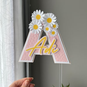 May include: Pink cake topper with the letter "A" and the name "Adi" in yellow script. The letter is decorated with white daisies with yellow centers.