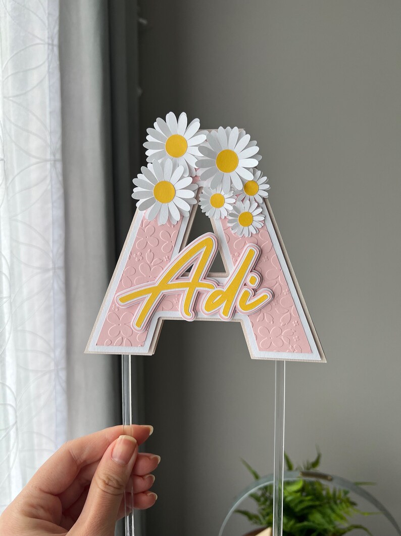May include: Pink cake topper with the letter "A" and the name "Adi" in yellow script. The letter is decorated with white daisies with yellow centers.