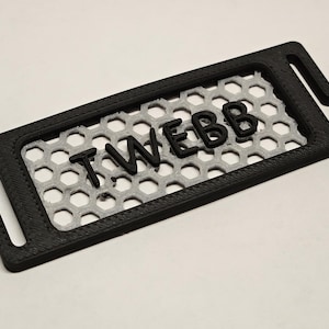 May include: A black and gray rectangular 3D-printed item with the text "T WEBB" in black. The item has a honeycomb pattern in the center and hooks on each end.