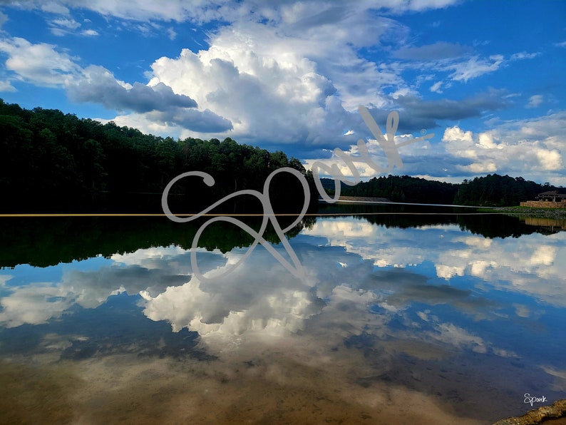 Peut inclure: Un lac paisible avec un reflet de nuages blancs et moelleux sur un ciel bleu. Le lac est entour&eacute; d'arbres verdoyants.