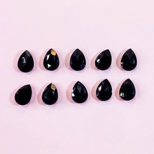 May include: Ten black teardrop-shaped gemstones arranged on a light pink background. Some of the gemstones have a gold-colored accent. The gemstones are faceted, reflecting light. The overall composition is simple and elegant.