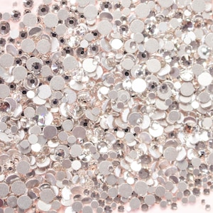May include: A collection of silver rhinestones of varying sizes, scattered across a light pink surface. The rhinestones are round and faceted, reflecting light. The image is a close-up, highlighting the sparkle and detail of the embellishments.
