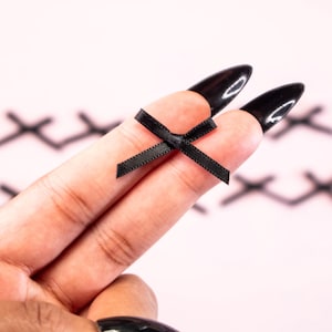 May include: A small black satin bow is held between two fingers with black nail polish. Several similar bows are blurred in the background on a pink surface. The bows are likely for crafts or embellishments.