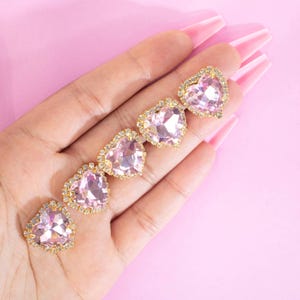 May include: Five pink heart-shaped rhinestones with gold settings. The rhinestones are arranged in a row on a pink surface.