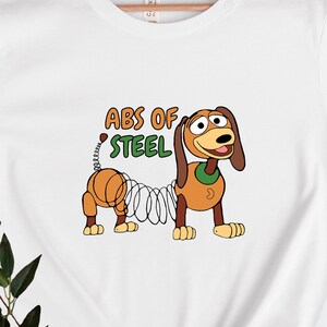 Può includere: Maglietta bianca con un'immagine di Slinky Dog di Toy Story in stile cartone animato. Il testo "ABS OF STEEL" è sopra l'immagine.