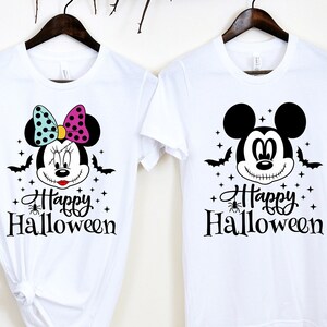 Puede incluir: Dos camisetas blancas con tem&aacute;tica de Halloween. Una camiseta presenta un Mickey Mouse en blanco y negro con el texto "Happy Halloween". La otra camiseta presenta una Minnie Mouse colorida con el texto "Happy Halloween".