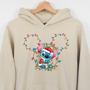 Może przedstawiać: Beżowa bluza z kapturem z niebieską postacią Stitch, kt&oacute;ra ma czapkę Świętego Mikołaja i jest opleciona świątecznymi lampkami. Lampki są ułożone w kształcie uszu Myszki Miki.