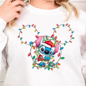 Może przedstawiać: Biała bluza z niebieską postacią Stitch, kt&oacute;ra ma na sobie czapkę Świętego Mikołaja i jest zaplątana w świąteczne lampki. Lampki mają kształt uszu Myszki Miki.